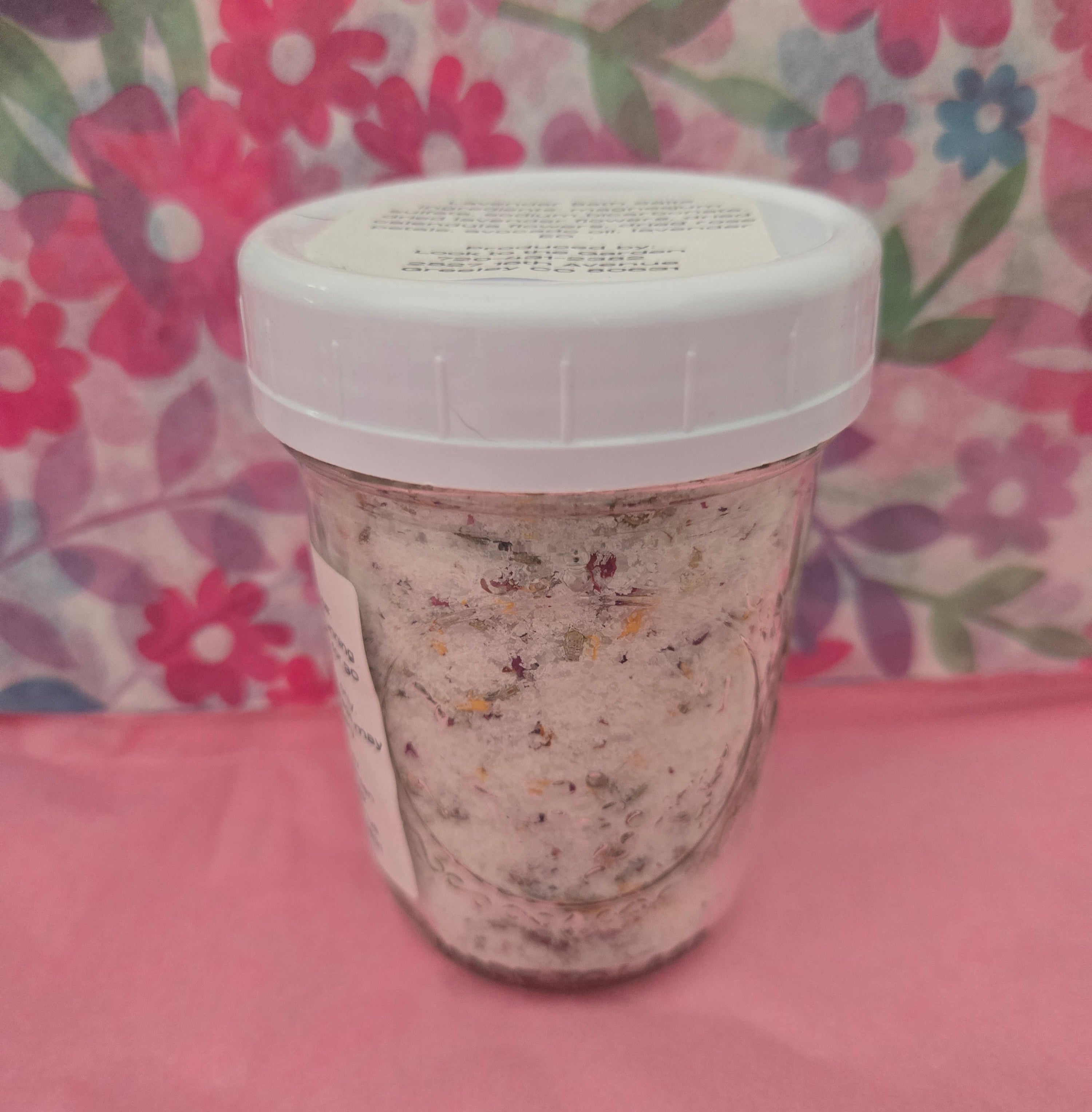 Clear jar containing lavender bath salts with a white lid on a pink surface with a floral background